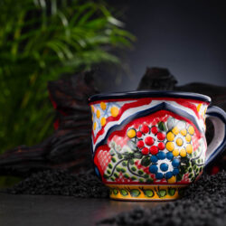 bunte Kaffeetasse rot-blau-gelb mit Blumen auf Schieferplatte mit Deko und Palmenblättern
